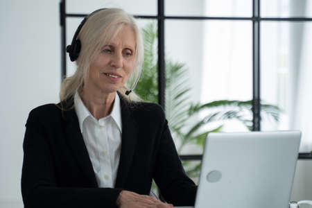 Elderly lady in a headset talks on a video conference, conducts a video conference using a laptop in the officeの写真素材