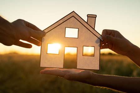 Family holds paper house at sunset, sun shines through window. Hand holding paper cut of house symbols at sunsetの写真素材