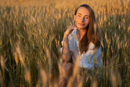 Young slender woman in a white shirt, sitting on a wheat field on a summer dayの写真素材