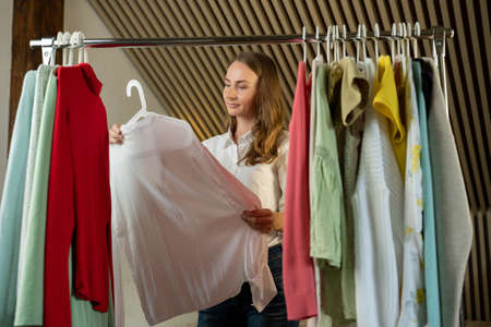 Beautiful thoughtful woman stylist chooses clothes on a hanger in the dressing roomの写真素材