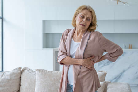 An elderly woman feels back pain while massaging aching muscles while standing in the living roomの写真素材
