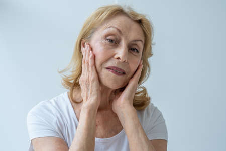 Close-up portrait of a happy elderly woman looking in the mirror, touching her skinの写真素材