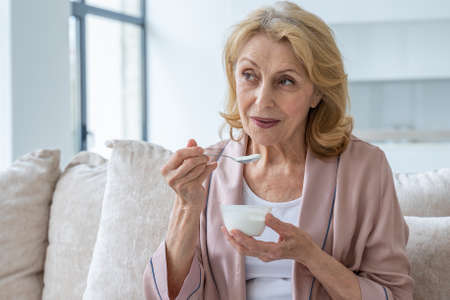 Smiling elderly woman who eats yogurt sitting on the couchの写真素材