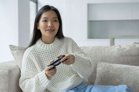 An Asian woman is sitting on the couch, holding a joystick and playing a game on the console. The...