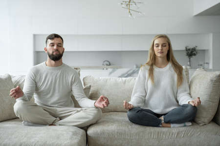 Calm young family is doing yoga together, sitting in the lotus position on the sofa. Parents relax on the couch, meditatingの写真素材