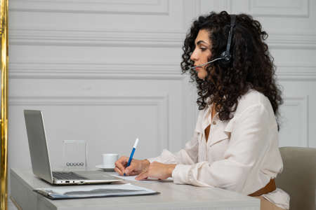 Side view smiling Indian employee with curly hair in headphones talks via video link, holds a web...