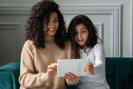Young black-haired curly-haired mother and daughter celebrate success with digital tablet, enjoy being home togetherの写真素材