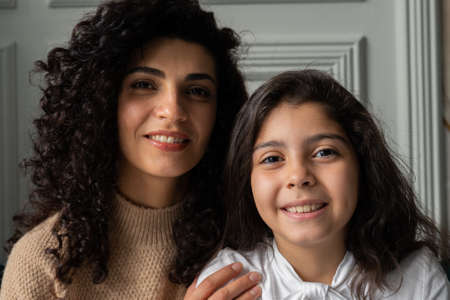 Young, curly-haired mother and her young daughter sit on the couch looking at the camera and smilingの写真素材