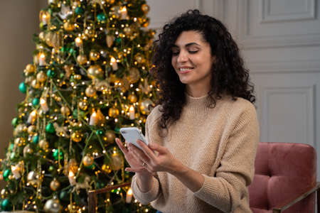 Smiling young woman using a smartphone, reading Christmas greetings on a social network, sitting on a sofa near a decorated Christmas tree.の写真素材