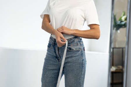 Slender black woman standing in front of a mirror measures her waist with a measuring tape after...