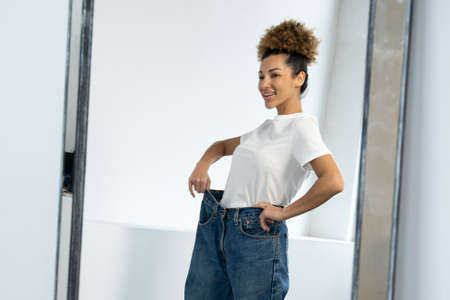 African-American woman dressed in big jeans before losing weight, looking at the figure in front of the mirror, is very glad that she has successfully lost weightの写真素材
