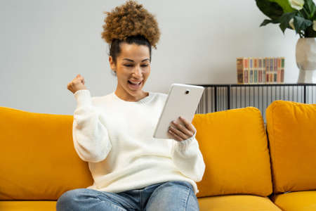 Excited African-American woman using a digital tablet, sitting at home on the couch, gets good news,...