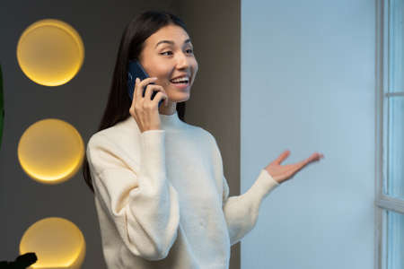 Young Asian woman is talking on a mobile phone and smiling, standing at the window, a freelance woman is calling on the phone, the concept of communication.の写真素材