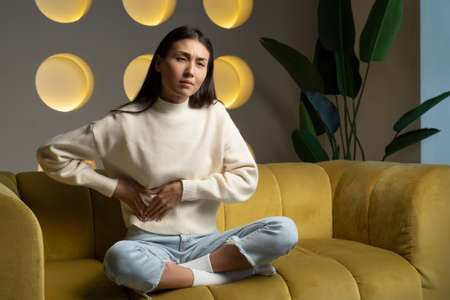 Young Asian woman in a sweatshirt, whose hands touch her stomach, suffers from abdominal pain while...