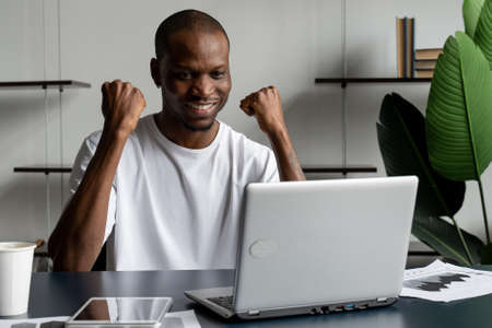 Happy excited businessman celebrates his success. The winner, a black man in the office reading the news on a laptopの写真素材