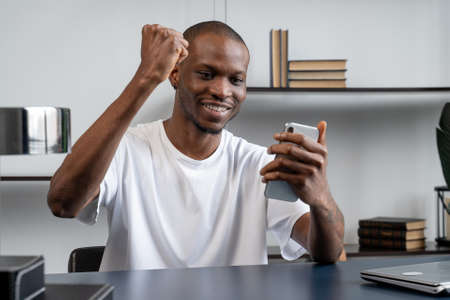 Black guy at the desk in the office is smiling happily, holding a smartphone in his hands. The delighted businessman looks at the smartphone screen and rejoicesの写真素材