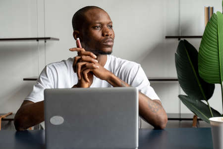 Thoughtful black businessman sitting at a desk with a computer and looking away. Distracted from his studies, the young man plunged into thoughtの写真素材