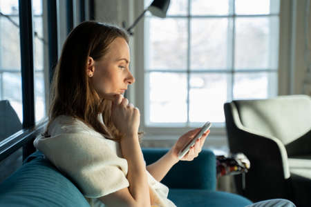 Serious excited young woman exchanges text messages using a mobile phone while sitting at home on the couch.の写真素材