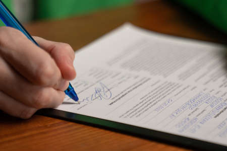Woman signs document with pen sitting at wooden table in office room. Female hand puts signature on...