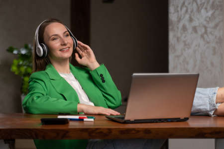 Cheerful woman enjoys music in headphones sitting at workplace near laptop. Young employee in green...