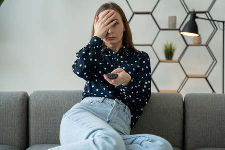 Close-up portrait of a young woman sitting on a sofa, watching TV, holding a remote control, shocked...