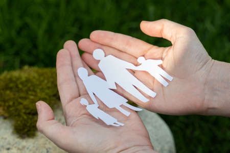 Paper origami as family image on woman palms on green garden blurred background closeup. Woman holds cut origami in hands as symbol of strong family relationshipsの写真素材