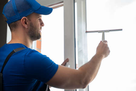 Worker in blue uniform cleans plastic window with detergent and squeegee. Soapy stains on window glass after using detergent. Windows cleaning in private houseの写真素材