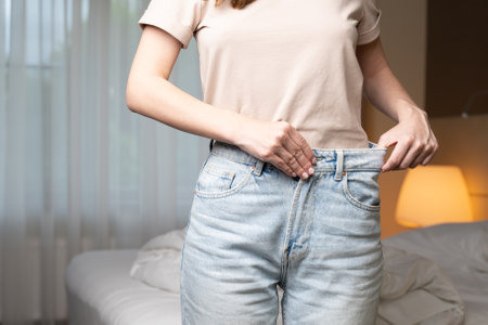 Woman tries on old jeans after loosing weight standing near bed in hotel room. Lady encourages to keep healthy diet showing fresh apple closeupの写真素材