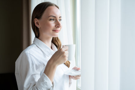 Happy young woman comes to window with white curtain drinking cup of coffee. Female guest enjoys...