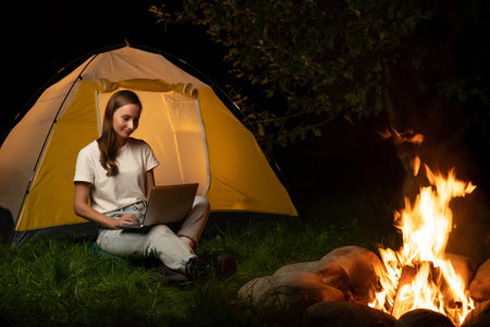 Young freelance woman working on a laptop in the evening. A female tourist is sitting by a campfire and a tentの写真素材