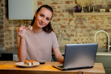 Young pretty woman sitting at her kitchen eat sushi at table home working with a laptop.の写真素材