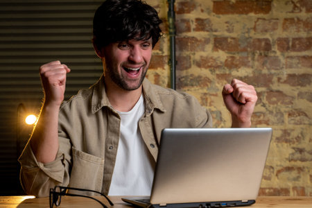 Happy businessman with glasses, using a laptop at work, raises his fists, celebrating good results. An excited man looks at the computer, enjoying playing the lottery jackpotの写真素材