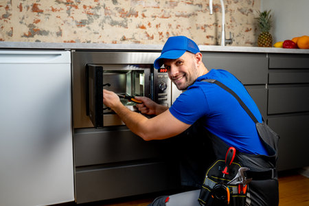 Young repairman in a blue uniform repairs and repairs a microwave oven with a screwdriver in a modern kitchen.の写真素材