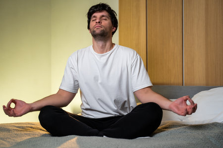 Positive guy in a white T-shirt and black pants, sitting on the bed in the bedroom, doing yoga in...