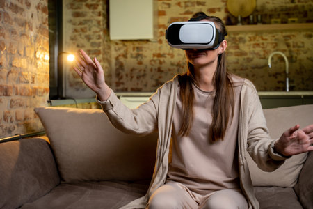 Young woman sitting on a sofa in her own living room wearing virtual reality glasses is browsing an...