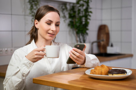 Happy woman who eats breakfast and uses a smartphone. The girl laughs, eats toast and drinks coffee, holding a mobile phone in the kitchen at home.の写真素材