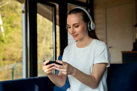 Beautiful woman is playing a mobile game and wearing wireless headphones while sitting on a blue...