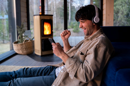 Young man is sitting on the floor by the sofa with headphones and a phone in his hands against the background of the fireplace. Listens to music, podcast.の写真素材