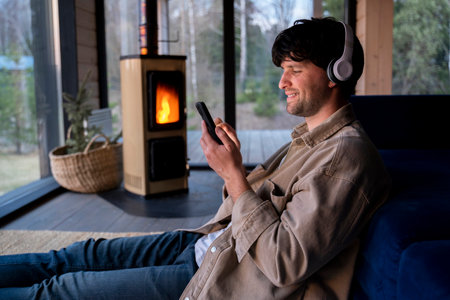 Young man is sitting on the floor by the sofa with headphones and a phone in his hands against the background of the fireplace. Listens to music, podcast.の写真素材
