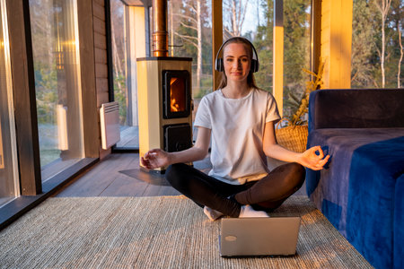 Young woman wearing headphones during an online yoga meditation practice. A woman sits in a lotus position in front of a laptop monitor and meditates with her eyes closed in the living room against the background of a fireplace.の写真素材