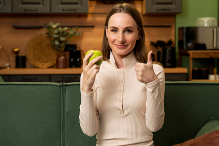 Young brunette woman eats a green apple sitting on a green sofa in the living room.の写真素材