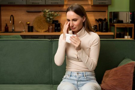 Young brunette woman is crying, sitting alone on a green sofa at home in the living room.の写真素材