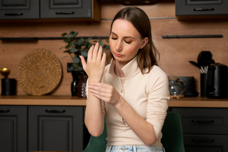 A young brunette woman touches a sore wrist while sitting at a table in the kitchen at home.の写真素材