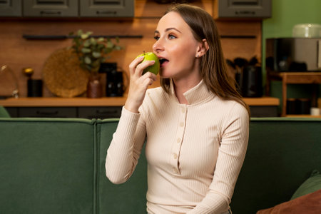 Young brunette woman eats a green apple sitting on a green sofa in the living room.の写真素材