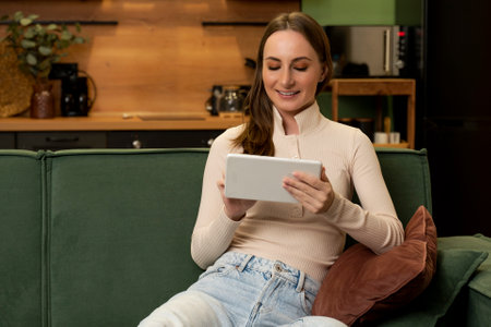 A young charming smiling Caucasian brunette in a beige blouse is sitting on a green sofa in the living room using a tablet.の写真素材