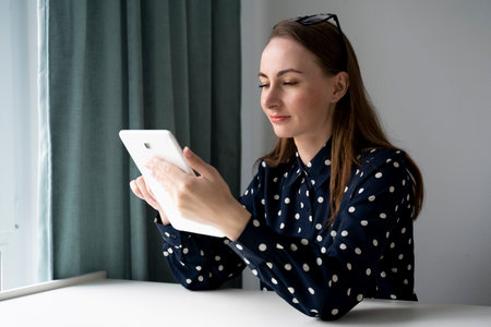 Freelance asian woman surfing browser with tablet sitting with pillow on sofa at living room. Beautiful teenager girl using digital tablet enjoy shopping online or watching ebook at homeの写真素材