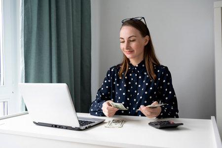 A young woman in a shirt, counting and handing out cash to pay bills at a table, using a laptop.の写真素材