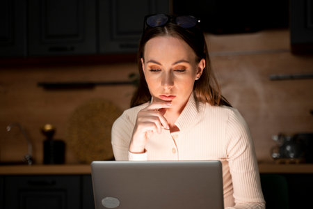 A young brunette woman makes purchases on the Internet, uses a laptop in the evening in the kitchen.の写真素材