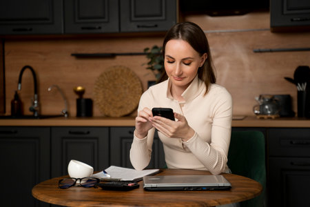Woman sitting at the kitchen table with a mobile phone is using and smiling.の写真素材