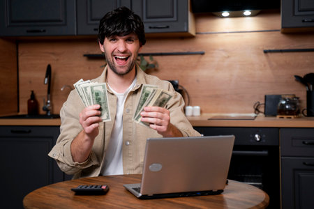 A smiling businessman calculates the monetary income, holding dollar bills in his hands. A man uses a calculator while paying for utilities in the kitchen.の写真素材